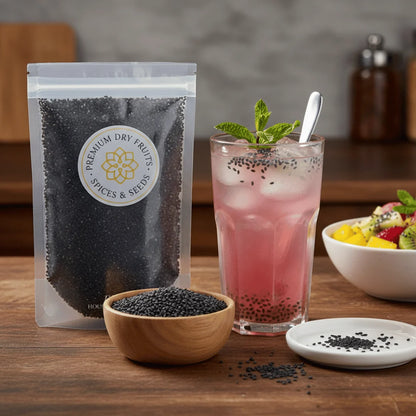 Packaging of premium Basil Seeds, a bowl of seeds, a glass of pink drink with ice and mint leaves, and a bowl of fruit salad on a wooden table.