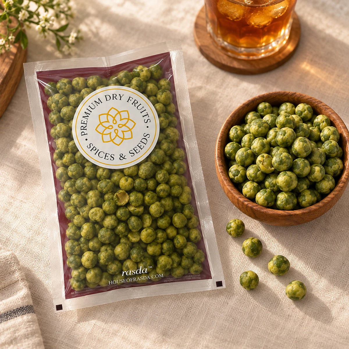 Wasabi coated green peas in a pouch on a linen surface with a wooden bowl of peas and a glass of iced tea